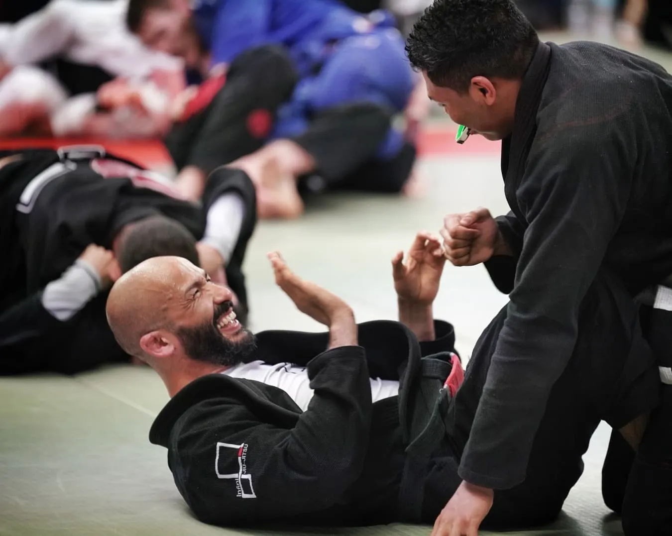 Un pratiquant en kimono noir éclate de rire au sol pendant l’entraînement